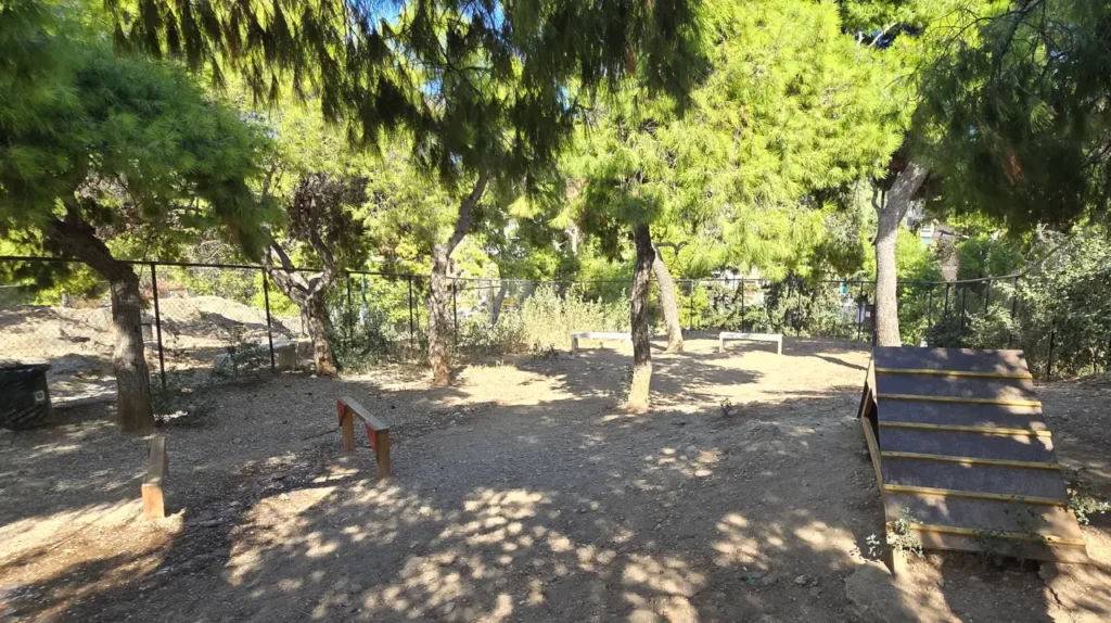Ευρύχωρος περιφραγμένος χώρος με πεύκα, ξύλινη ράμπα agility και εμπόδια στο πάρκο σκύλων στον Λόφο Λαμπράκη